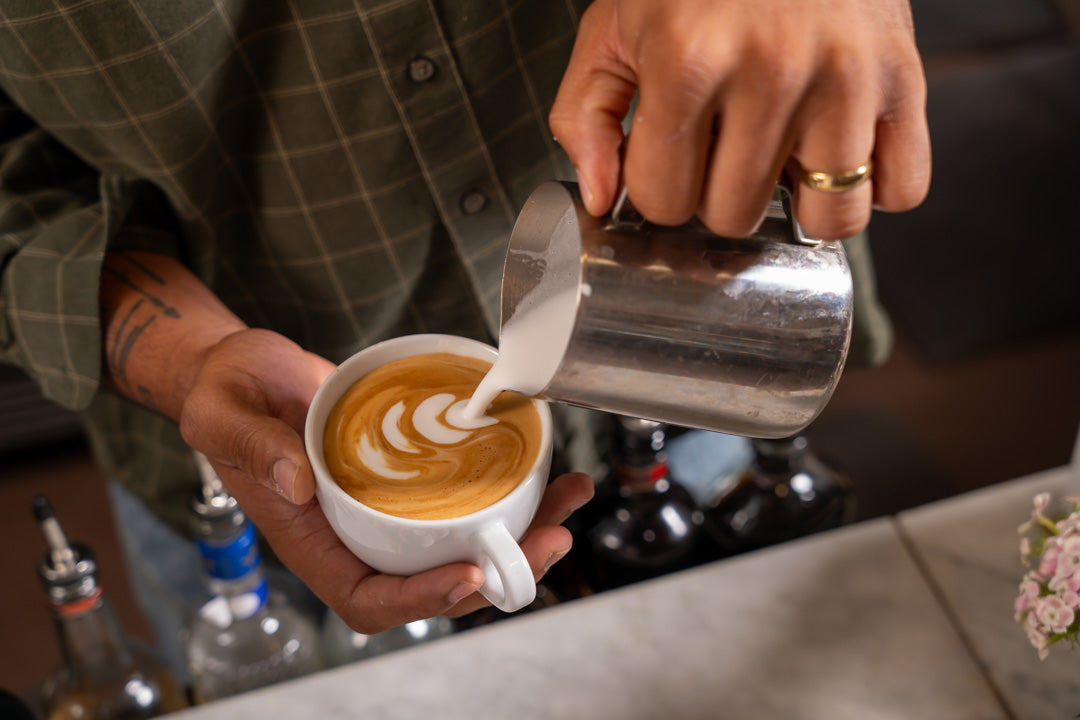 A barista carefully pours steamed milk into a white ceramic cup to create latte art.