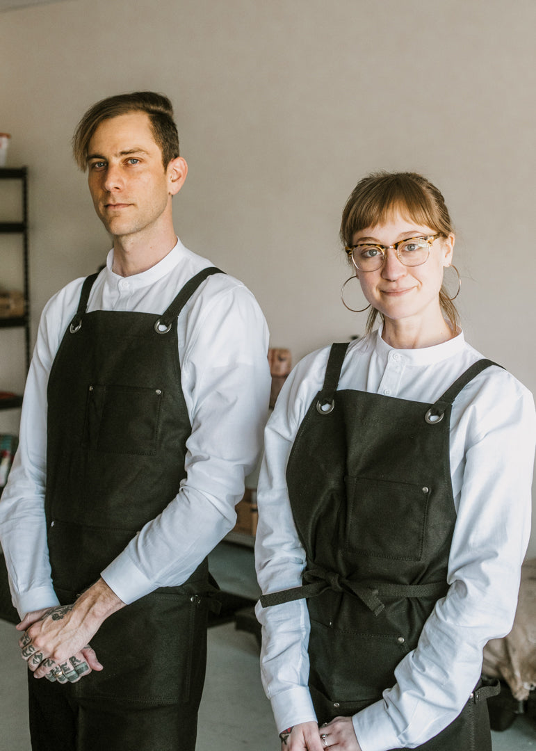 Christopher Pierce and Ashley Griffeth stand for a photo, wearing matching black aprons and white undershirts in the Loom Coffee Co. roasting facility.