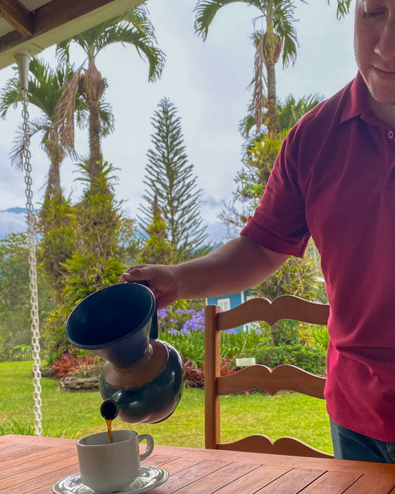 Aquiares Estate resident pouring freshly brewed coffee from a vandola against a backdrop of palm trees and tropical foliage.